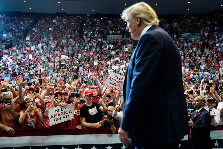A woman in camouflage raises her hand while holding a sign that reads "I Stand With President Donald J. Trump," surrounded by Trump flags and supporters.
